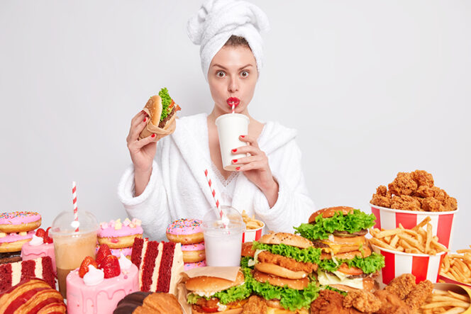 Unhealthy nuttrition concept. Housewife with red manicure and lips in domestic bathrobe towel on head drinks soda eats hamburger eats delicious fast food stands against white background. Breaking diet