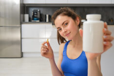 Close up portrait of young woman, fitness instructor showing bottle of vitamins, taking buds supplementary, dietary pill, sitting at home, doing workout exercises